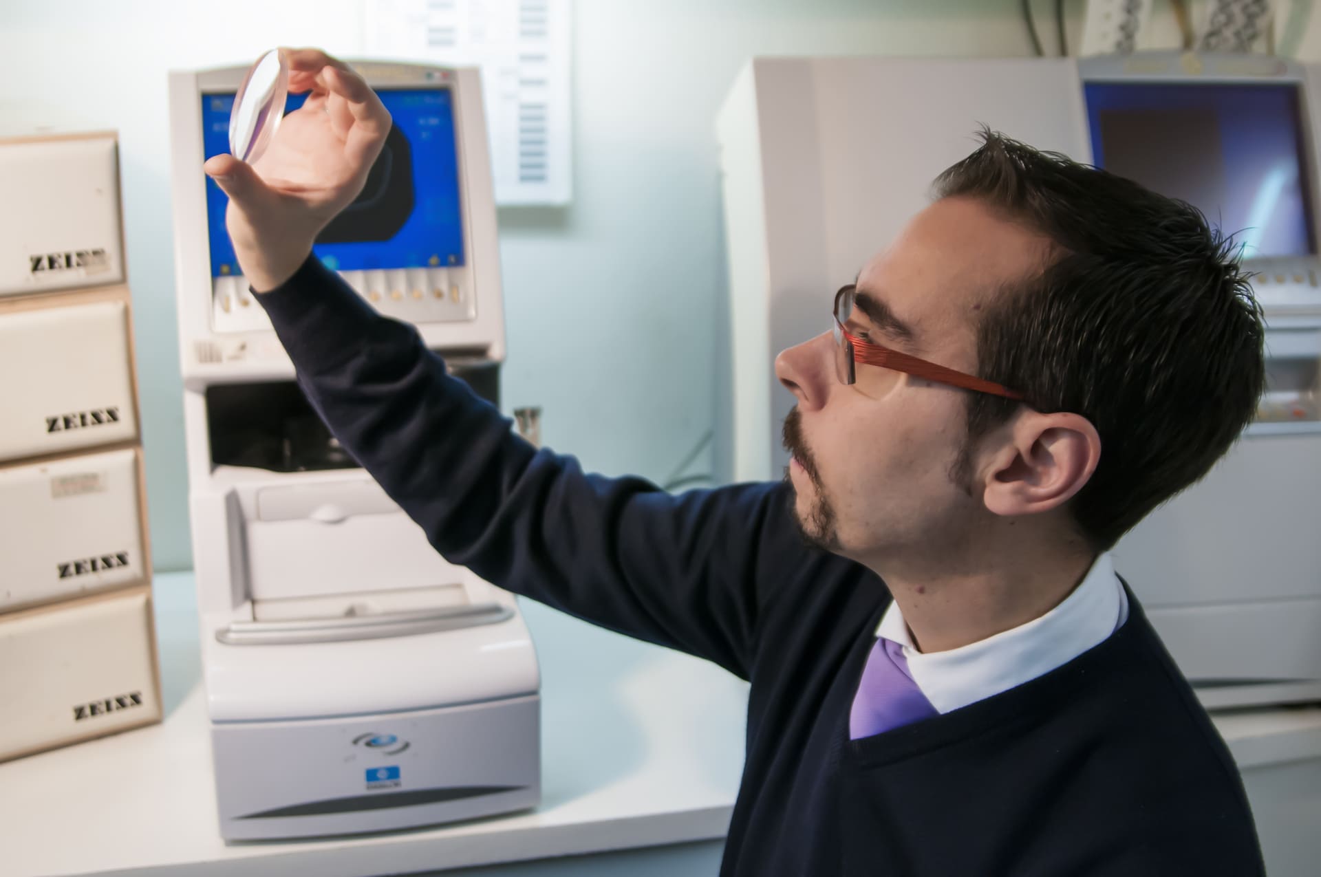 Un uomo con maglione scuro e occhiali da vista solleva una lente ottica per esaminarla - Crisafulli Eyewear a Milano