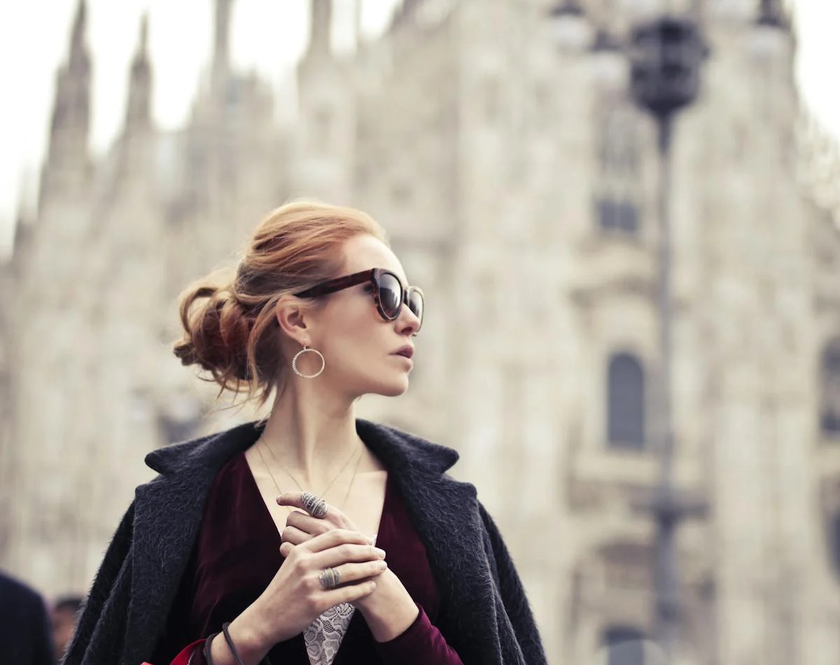 A woman with red hair, sunglasses, and a dark coat, holds her hands together - Crisafulli Eyewear in Milan